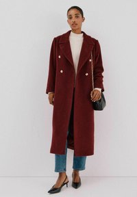 Lange bordeauxrode jas met een dubbele knoopsluiting en metalen knopen, gecombineerd met een crèmekleurige trui, blauwe jeans en zwarte slingback schoenen.