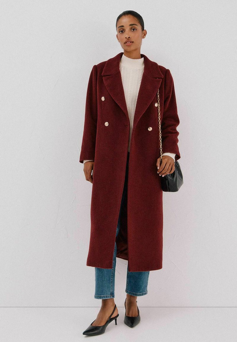 Lange bordeauxrode jas met een dubbele knoopsluiting en metalen knopen, gecombineerd met een crèmekleurige trui, blauwe jeans en zwarte slingback schoenen.