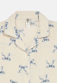 Ljusbeige pyjamasöverdel med blå ripsband och rosetttryck. Har en vikkrage, knappstängning och kontrasterande passpoal längs kanterna.