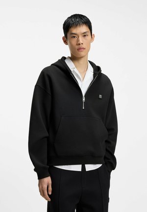 Ung mand iført en sort hoodie med halv lynlås over en hvid skjorte og sorte bukser, stående mod en ensfarvet hvid baggrund.