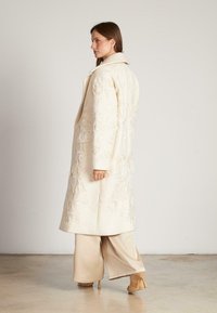Abrigo largo color crema con intrincados bordados florales, cuello con muesca y tejido texturizado, combinado con pantalones beige de pierna ancha y tacones.