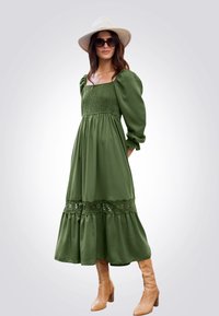 Abito lungo verde a maniche lunghe con corpino arricciato, gonna a strati e dettagli in pizzo all'orlo. Indossato con stivali beige sopra il ginocchio e cappello a tesa larga.