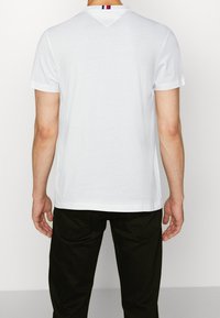 T-shirt bianco a maniche corte con scollatura a V, caratterizzato da un dettaglio a righe sul collo posteriore. Tessuto morbido con una texture liscia.