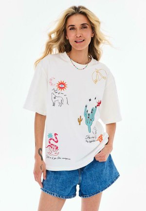 Kobieta z długimi blond włosami, ubrana w biały t-shirt z kolorowymi mistycznymi i nawiązującymi do natury nadrukami oraz niebieskie jeansowe szorty.