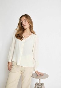 Jasno-kremowa bluzka z długimi rękawami, z miękkiego materiału, z okrągłym dekoltem. Połączona z beżowymi spodniami, prezentowana na jednolitym tle.