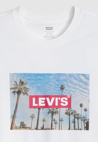 Tricou alb din bumbac, cu un grafic colorat de palmieri pe un fond albastru, având un logo Levi's roșu, proeminent, în centrul imaginii.
