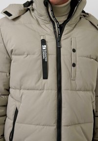 Bežinis pūkinis švarkas su užtrauktuku priekyje, dviem užtrauktukais uždaromomis kišenėmis ir gobtuvu. Savybės: juodi akcentai ir audinio etiketė ant krūtinės.