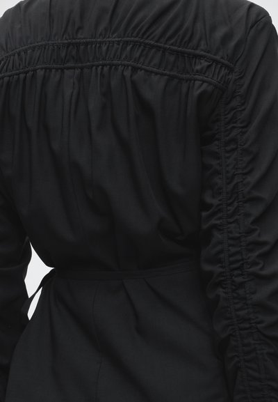 Dos d’un vêtement noir avec des détails élastiques froncés sur les épaules et le long des manches, avec une ceinture à nouer à la taille.