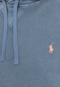 Sininen huppari, johon on ommeltu kiristysohjaimet ja pieni pinkki brodeerattu hevosen ja ratsastajan logo rinnassa.