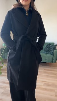 Juodas apgaubtas paltas su ilgomis rankovėmis, dirželiu juosmens srityje ir dideliais atvartais. Pagamintas iš minkštos, tekstūruotos medžiagos. Kišenės kiekvienoje pusėje.