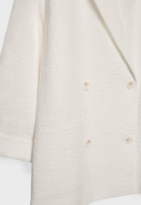 Blazer croisé de couleur crème en tissu texturé, avec quatre boutons et une poche passepoilée unique sur le côté gauche.