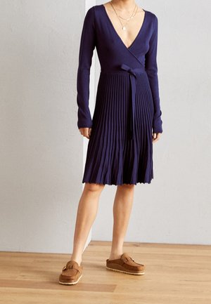 Femme portant une robe portefeuille plissée à manches longues bleu marine avec ceinture nouée à la taille et des chaussures marron sans lacets, debout sur un sol en bois contre un mur clair.