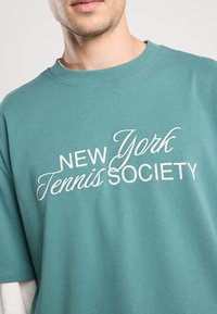 Turkoosi puuvillapaita, jossa on valkoinen brodeerattu teksti "New York Tennis Society." Pyöreä kaula-aukko ja lyhyet hihat, sileä pinta.