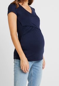 Gravid kvinna som bär en marinblå kortärmad graviditetstopp och ljusblå jeans, stående mot en vit bakgrund.
