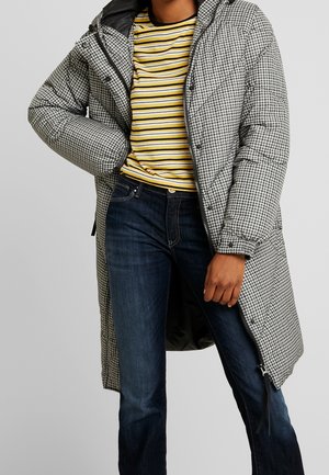 Personne portant un long manteau matelassé à motif pied-de-poule par-dessus une chemise rayée jaune, noire et blanche, avec un jean bleu marine, devant un fond gris.