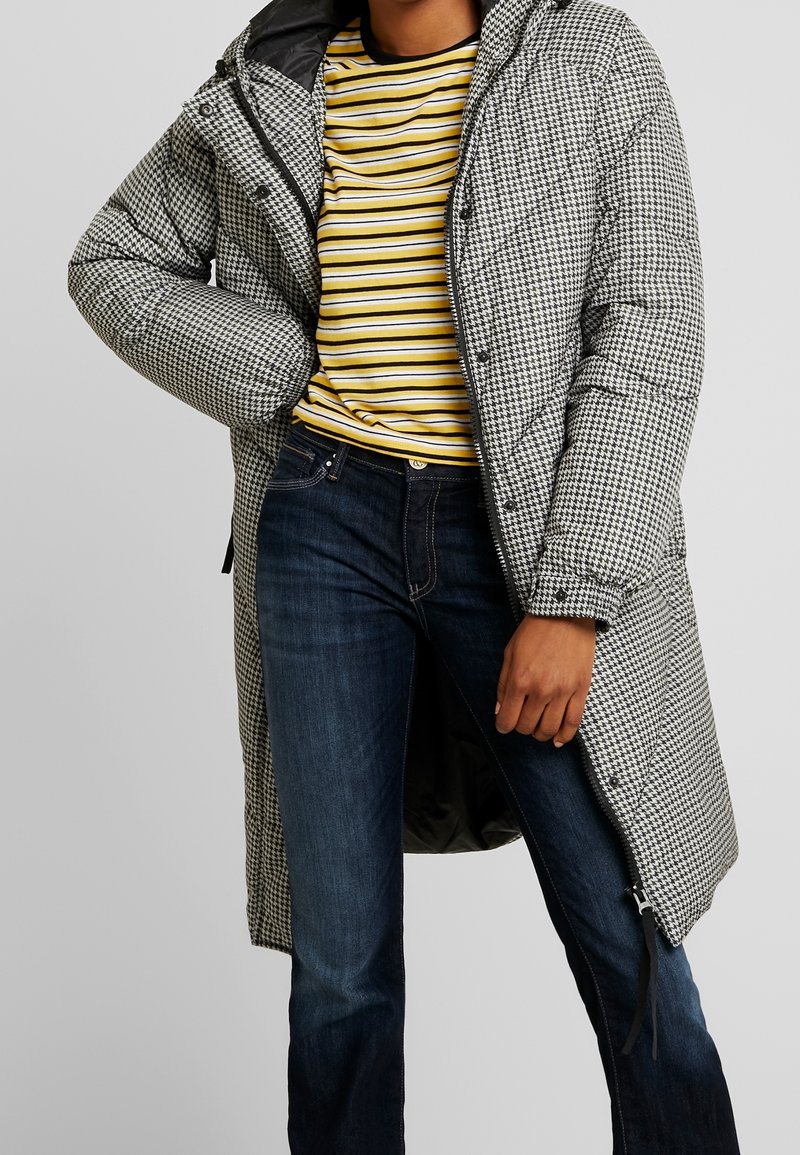 Personne portant un long manteau matelassé à motif pied-de-poule par-dessus une chemise rayée jaune, noire et blanche, avec un jean bleu marine, devant un fond gris.