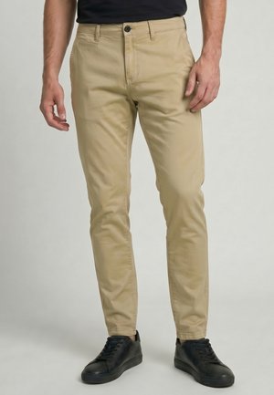 Homme portant un pantalon beige slim-fit et des baskets noires, debout devant un fond clair et neutre.