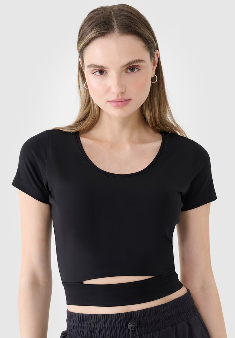 Jeune femme aux longs cheveux bruns portant des boucles d'oreilles argentées en forme d'anneau, un crop top noir à manches courtes avec une découpe et un pantalon noir froncé.