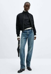 Schwarze Bomberjacke über einem dunklen Pullover, blaue Jeans und schwarze Knöchelboots. Das Model hält eine schwarze Handtasche, mit strukturiertem Haar, das in Zöpfen frisiert ist.