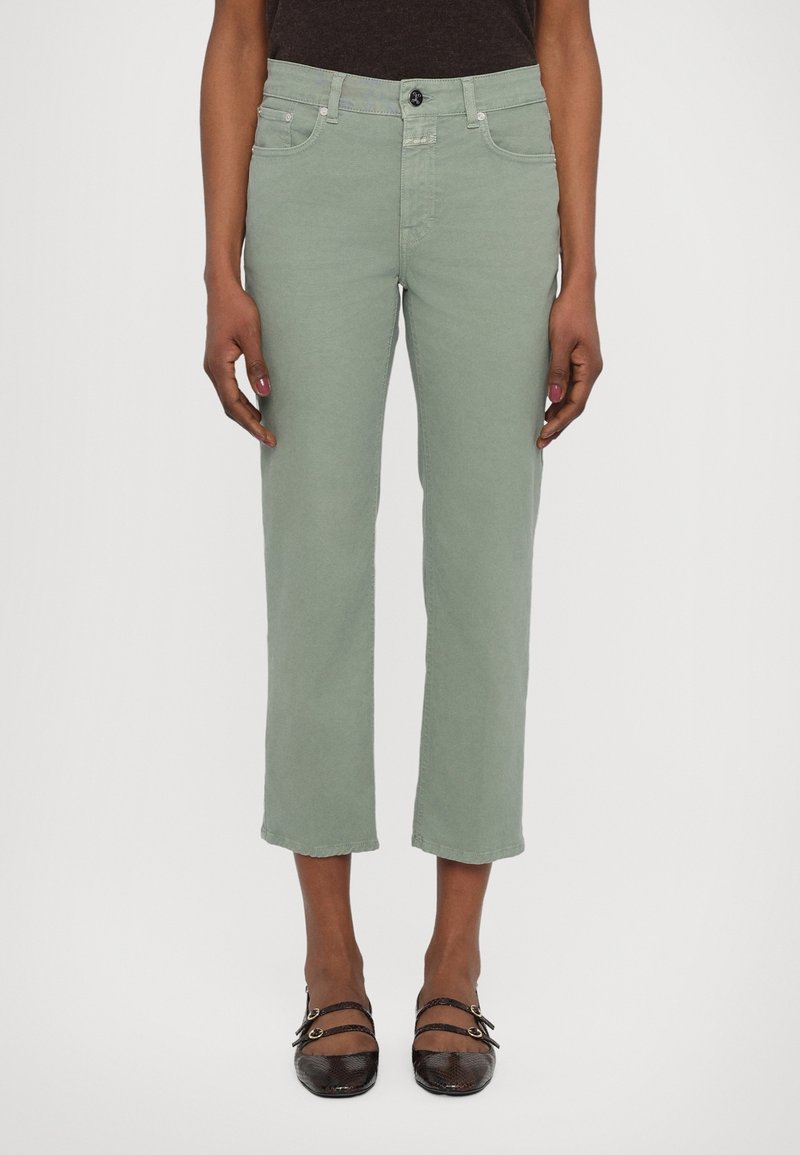 Femme portant un pantalon vert clair court à jambes droites et des chaussures noires à double bride, debout devant un fond uni.