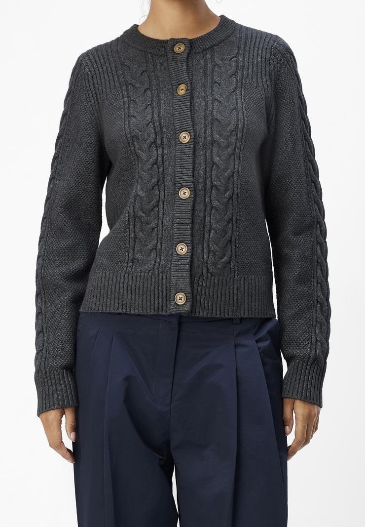 Femme portant un cardigan boutonné gris foncé en maille torsadée avec des boutons en bois et un pantalon plissé bleu marine.
