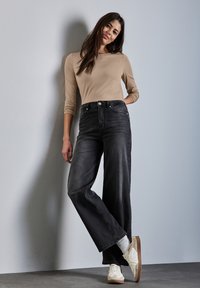 Model trägt ein enganliegendes, langärmliges beige Oberteil und hochgeschnittene schwarze weit geschnittene Jeans, kombiniert mit weißen Sneakers und grauen Socken.