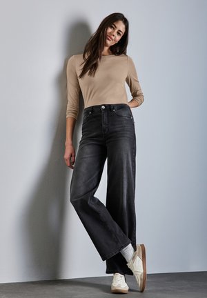 Modèle portant un haut beige ajusté à manches longues et un jean noir large taille haute, associé à des baskets blanches et des chaussettes grises.