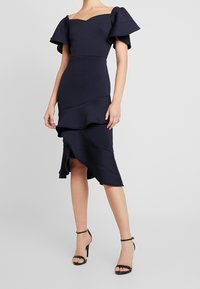 Marinblå av-axel-klänning med volangärmar och lagerstruktur i nederkant. Tillverkad av stretchtyg, med en figurnära silhuett och utvidgande detaljer.