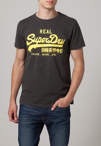 Camiseta de manga corta gris con estampado amarillo que dice "REAL SuperDry", con un cuello redondo clásico y un corte relajado.