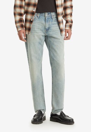 Mann trägt hellblaue Jeans mit geradem Bein, schwarze Slipper mit weißen Socken, ein schwarzes Hemd und ein aufgeknöpftes braunes karierte Hemd.