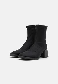 Bottines noires en tissu doux et extensible. Elles présentent un bout pointu et un talon épais, ainsi qu'une fermeture éclair à l'avant.