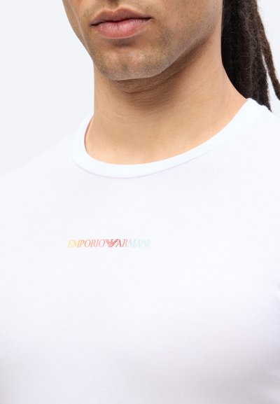 Κοντινό πλάνο ενός ατόμου που φοράει λευκό T-shirt με μικρό, πολύχρωμο λογότυπο "Emporio Armani" στο στήθος.