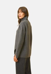 Grauer Strickpullover mit hohem Kragen, geripptem Textur, seitlichen Schlitzen und lockerer Passform. Getragen mit dunklen Hosen.