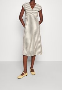 Bezramenné béžové šaty s vertikálními hnědými pruhy, výstřih do V a uvolněný střih, doplněné žlutými balerínami Mary Jane.