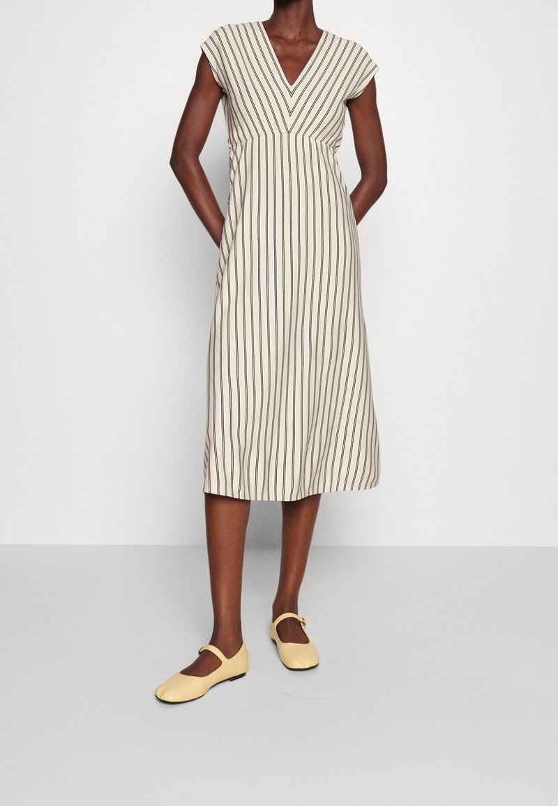 Bezramenné béžové šaty s vertikálními hnědými pruhy, výstřih do V a uvolněný střih, doplněné žlutými balerínami Mary Jane.