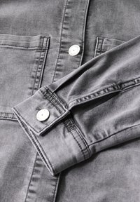 Camicia in denim grigio con bottoni a pressione argentati, due tasche sul petto e dettagli di cucitura visibili. Tessuto liscio e design casual.
