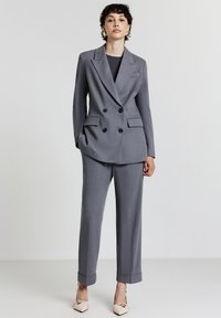 Persona che indossa un blazer grigio doppiopetto e pantaloni coordinati con risvolto, abbinati a décolleté beige a punta, in piedi di fronte a uno sfondo semplice.