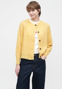 Mlada osoba s kratkom kosom koja nosi žutu kardigan sa dugmadi preko bijele majice i tamnoplavih traperica, stoji s jednom rukom u džepu.