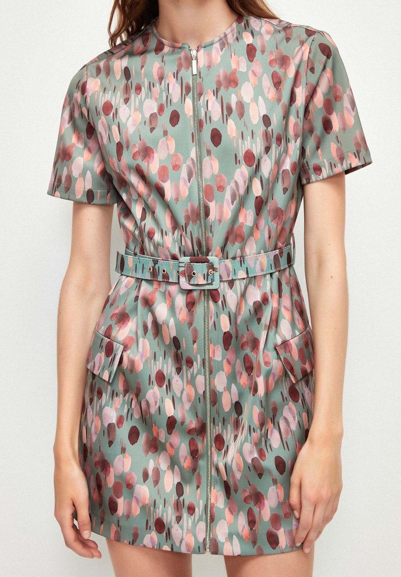 Robe à manches courtes en tissu vert avec un motif de points roses et bordeaux, équipée d'une fermeture éclair à l'avant et d'une ceinture assortie avec des poches.
