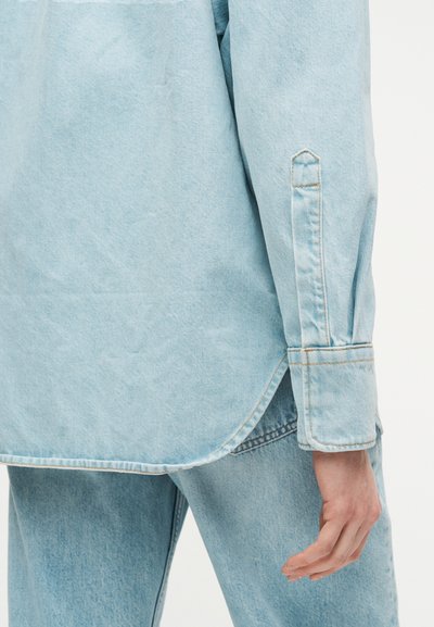 Μανσέτα μπλε πουκαμίσου από τζιν και πίσω τελείωμα πάνω από τζιν ίδιου χρώματος, που δείχνει ραμμένες λεπτομέρειες και χαλαρή εφαρμογή στην περιοχή του καρπού και του ισχίου.