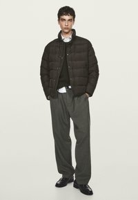 Giubbotto puffer marrone con chiusura a zip e bottone, indossato sopra un maglione scuro e una camicia bianca. I pantaloni grigi e le scarpe nere completano il look.