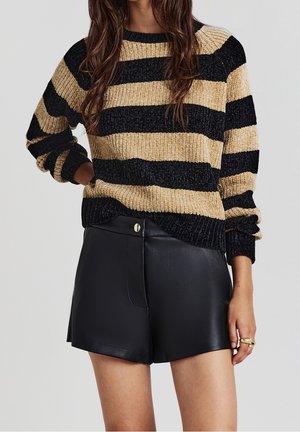 Femme portant un pull épais beige et noir à rayures, manches longues, et un short noir en similicuir taille haute.