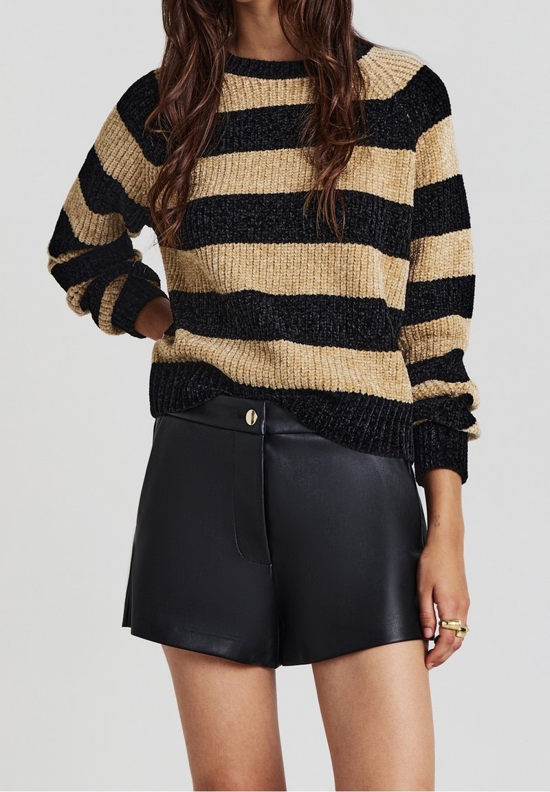 Femme portant un pull épais beige et noir à rayures, manches longues, et un short noir en similicuir taille haute.