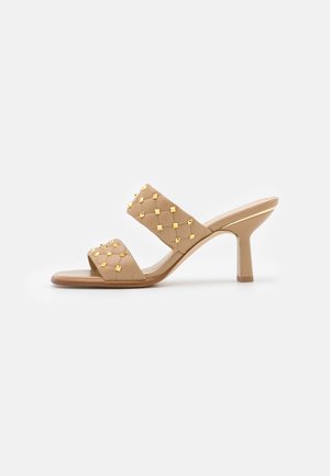 Sandale à talon beige avec deux brides matelassées ornées de clous carrés dorés et un talon fin de hauteur moyenne.