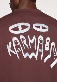 Camicia corta rossa con grafica bianca grande che presenta occhi e la parola "KARMA" stilizzata con illustrazioni a mano e linee ondulate.