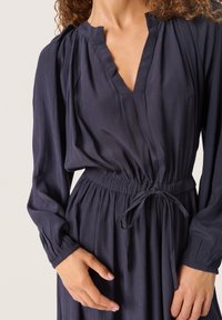 Langärmliges, dunkelblaues Kleid mit V-Ausschnitt, gerafften Schultern und Kordelzug in der Taille. Der Stoff hat eine weiche, strukturiert Oberfläche.