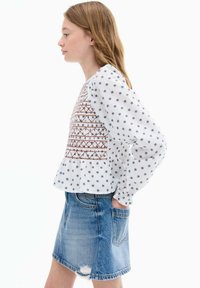 Bela bluzica z vezenimi detajli, dolgi napihnjeni rokavi in peplum rob, v kombinaciji z modrimi denim krili s sprednjim žepom in raztrganim robom.