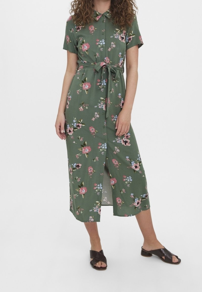 Femme portant une robe fleurie verte à manches courtes, boutonnée, avec une ceinture nouée à la taille et des sandales ouvertes à talons noires.