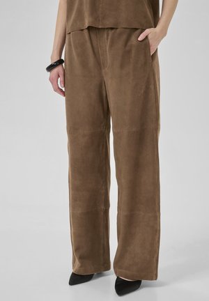 Una persona indossa pantaloni larghi in suede marrone con tasche laterali e scarpe nere a punta, abbinati a una maglietta coordinata in un ambiente neutro.