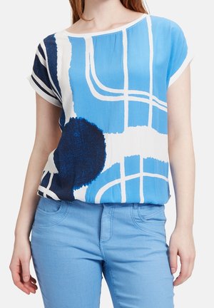 Vrouw draagt een kort mouw blauw-wit abstract patroon shirt met lichtblauwe broek, staand tegen een effen achtergrond.